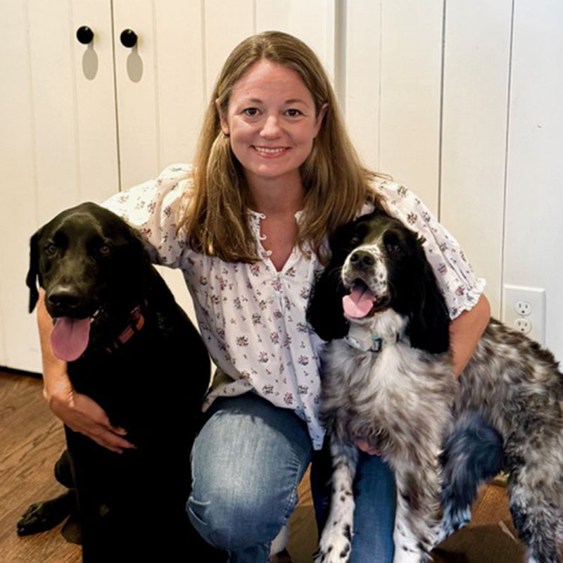 A woman kneels on a wooden floor, smiling between two dogs fresh from dog grooming. The dog to her left is large and black, while the dog to her right is medium-sized with a black and white coat. Both dogs have their tongues out, clearly enjoying their pampered state.