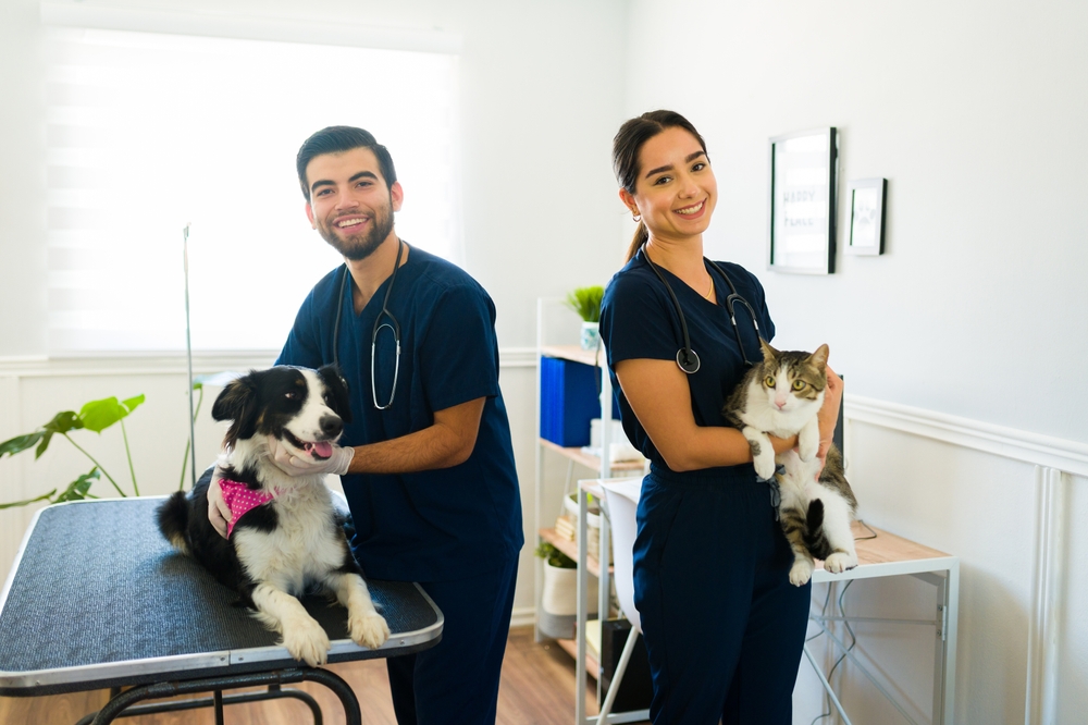 Two veterinarians in blue scrubs smile at the camera; one holds a cat while the other stands beside a dog on an exam table in a bright, modern clinic.